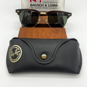 May include: Sunglasses with a dark brown frame and green lenses, resting on a wooden stand. A black leather case with a gold button and the Ray-Ban logo is in front. The text "SUNGLASSES BY BAUSCH & LOMB" is visible.