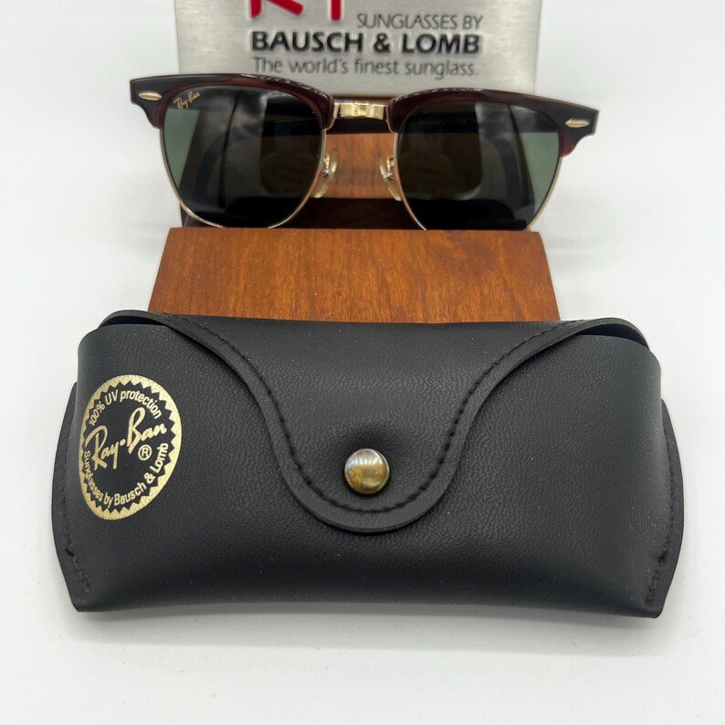 May include: Sunglasses with a dark brown frame and green lenses, resting on a wooden stand. A black leather case with a gold button and the Ray-Ban logo is in front. The text "SUNGLASSES BY BAUSCH & LOMB" is visible.