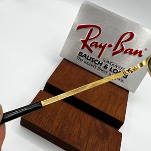 Puede incluir: Primer plano de gafas de sol Ray-Ban con montura dorada y patillas negras. Las gafas de sol descansan sobre un expositor de madera. Un letrero plateado con el logotipo y el texto de Ray-Ban est&aacute; detr&aacute;s de las gafas.