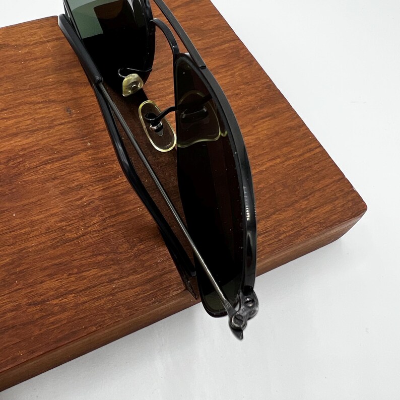 K&ouml;nnte beinhalten: Schwarze Sonnenbrille mit dunklen Gl&auml;sern und einem d&uuml;nnen Metallrahmen. Die Sonnenbrille liegt auf einer Holzoberfl&auml;che. Die Gl&auml;ser reflektieren die Umgebung.
