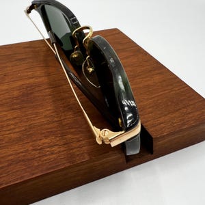 Pu&ograve; includere: Occhiali da sole neri con dettagli dorati esposti su un supporto in legno. Gli occhiali da sole hanno lenti scure e un design della montatura classico. Il supporto &egrave; di un ricco colore marrone scuro.