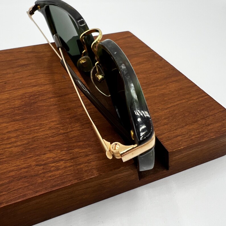 Pu&ograve; includere: Occhiali da sole neri con dettagli dorati esposti su un supporto in legno. Gli occhiali da sole hanno lenti scure e un design della montatura classico. Il supporto &egrave; di un ricco colore marrone scuro.