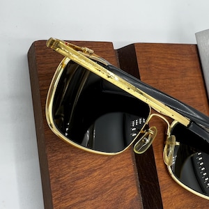 Puede incluir: Gafas de sol con montura dorada y lentes oscuras. Las gafas tienen forma rectangular y puente y patillas doradas. La parte superior de la montura es negra. Las gafas est&aacute;n sobre una superficie de madera.