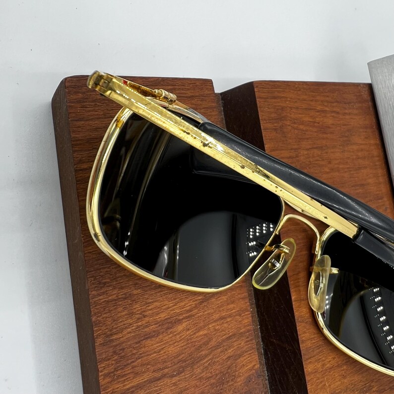 Puede incluir: Gafas de sol con montura dorada y lentes oscuras. Las gafas tienen forma rectangular y puente y patillas doradas. La parte superior de la montura es negra. Las gafas est&aacute;n sobre una superficie de madera.