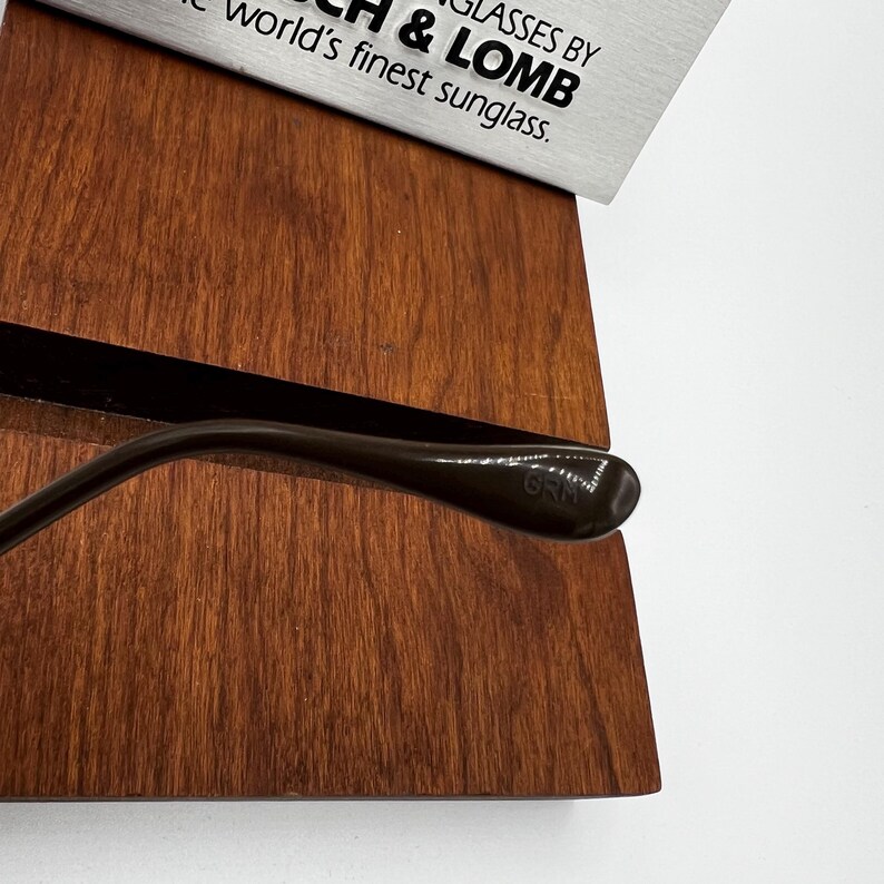 Pu&ograve; includere: Primo piano di un'asta di occhiali da sole marrone appoggiata su una superficie di legno. L'asta ha un design curvo e il testo "GRM" &egrave; visibile. La parte superiore dell'immagine mostra il testo "GLASSES BY B & LOMB" e "world's finest sunglass."