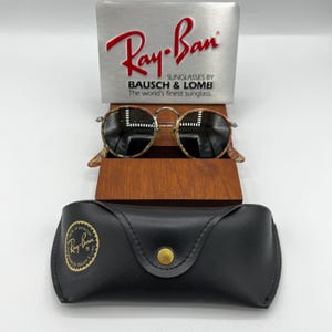 以下が含まれることがあります： べっ甲柄のフレームと濃い色のレンズが付いたRay-Banサングラス。木製のスタンドに展示されています。金色のボタンとRay-Banのロゴが付いた黒い革のケースが下にあります。ケースとディスプレイスタンドにRay-Banのロゴが見えます。