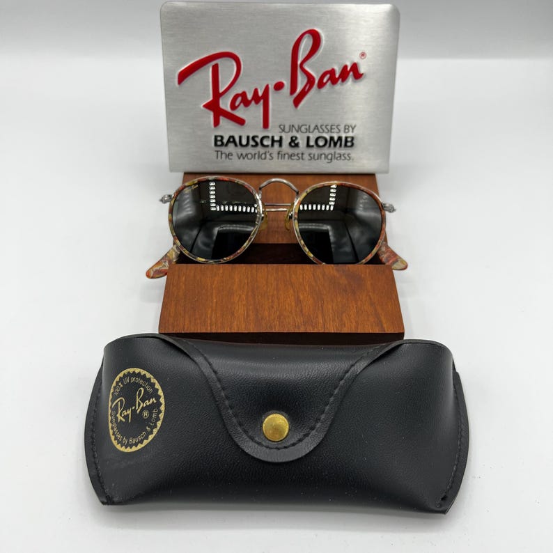 以下が含まれることがあります： べっ甲柄のフレームと濃い色のレンズが付いたRay-Banサングラス。木製のスタンドに展示されています。金色のボタンとRay-Banのロゴが付いた黒い革のケースが下にあります。ケースとディスプレイスタンドにRay-Banのロゴが見えます。