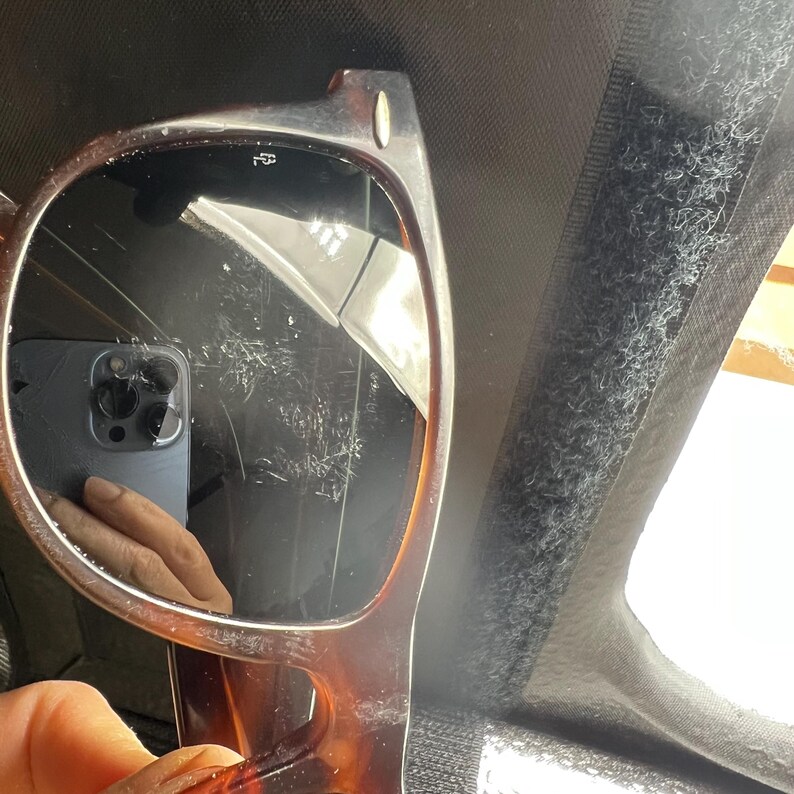 May include: Brown tortoiseshell sunglasses with dark lenses. The sunglasses have a classic rectangular frame design. The lenses reflect a phone and a hand. The sunglasses are held by a hand.