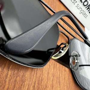 K&ouml;nnte beinhalten: Nahaufnahme einer schwarzen Sonnenbrille mit dunklen Gl&auml;sern und einem mattschwarzen Rahmen. Die Sonnenbrille hat einen klassischen Aviator-Stil mit einem Nasensteg aus Metall und transparenten Nasenpads. Das Bild ist vor einem Holzuntergrund aufgenommen.