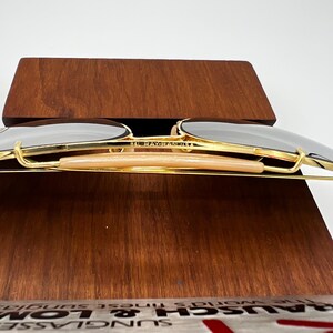May include: Gold-rimmed sunglasses with dark lenses, resting on a wooden display. The sunglasses have a classic aviator shape with a light-colored bridge and the text "BAUSCH & LOMB" is visible.