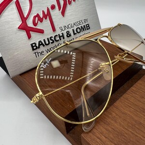 May include: Gold-tone aviator sunglasses with brown lenses. The sunglasses have a Ray-Ban logo on the left lens and the text "SUNGLASSES BY BAUSCH & LOMB The world's finest sunglasses" on the case.