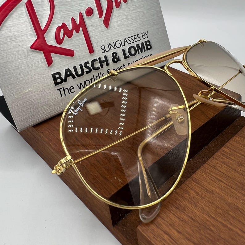 May include: Gold-tone aviator sunglasses with brown lenses. The sunglasses have a Ray-Ban logo on the left lens and the text "SUNGLASSES BY BAUSCH & LOMB The world's finest sunglasses" on the case.