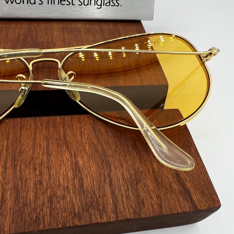 Puede incluir: Gafas de sol tipo aviador con montura de metal dorado y lentes tintadas en color &aacute;mbar. Las varillas tienen puntas de pl&aacute;stico transparente. Se ve un letrero plateado parcial en la parte superior con el texto "world's finest sunglass."