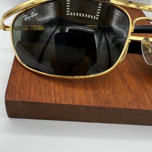 Puede incluir: Gafas de sol Ray-Ban con montura dorada y lentes oscuras. El logotipo de Ray-Ban es visible en la esquina superior izquierda de la lente. Las gafas de sol est&aacute;n sobre una superficie de madera oscura.