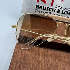 May include: Gold aviator sunglasses with brown lenses and a clear plastic nose bridge. The sunglasses are resting on a wooden surface.