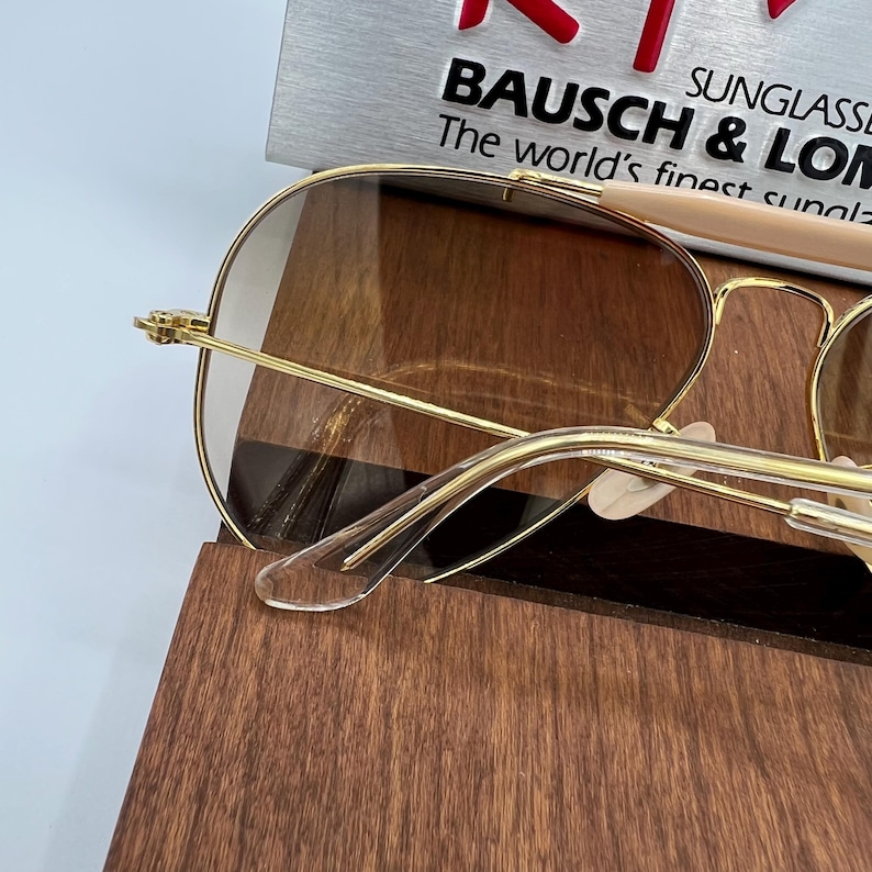May include: Gold aviator sunglasses with brown lenses and a clear plastic nose bridge. The sunglasses are resting on a wooden surface.