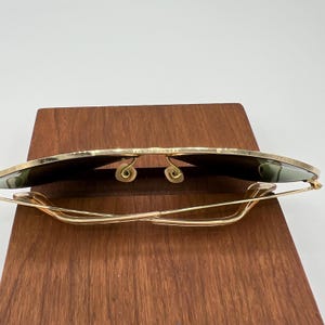 K&ouml;nnte beinhalten: Goldene Aviator-Sonnenbrille mit gr&uuml;nen Gl&auml;sern, die auf einer Holzoberfl&auml;che zusammengeklappt ist.
