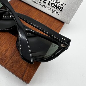 May include: Black sunglasses with a glossy finish, resting on a wooden surface. The sunglasses have a classic design with thick frames and dark lenses. The text on the background reads "GLASSES BY & LOMB world's finest sunglass."