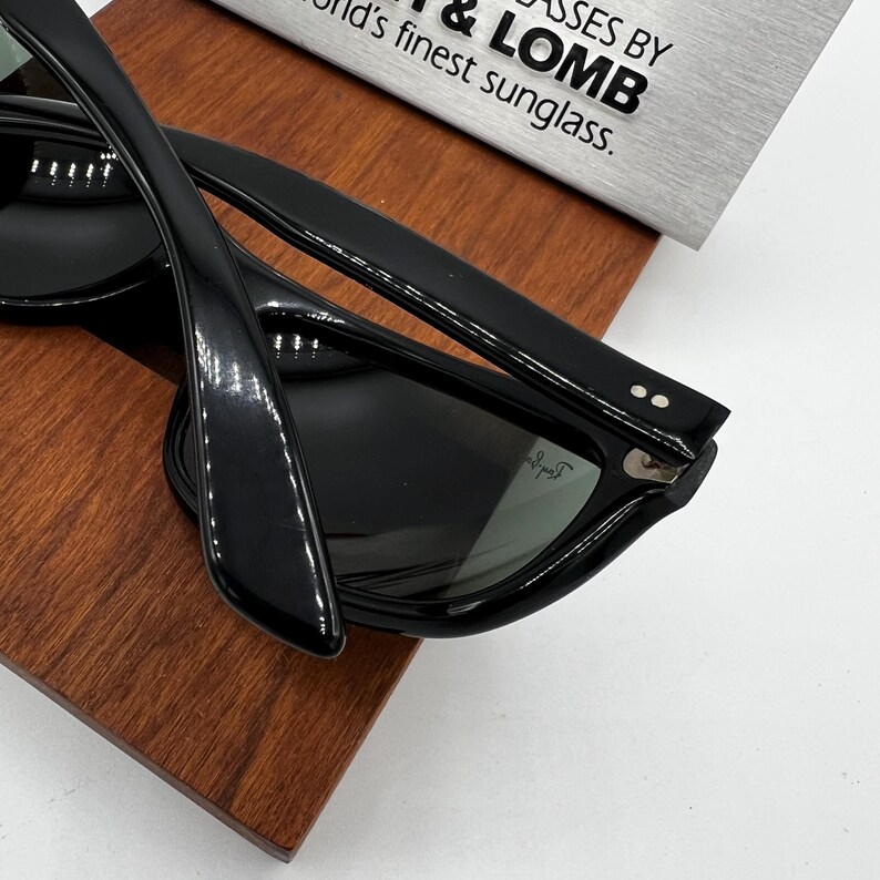 May include: Black sunglasses with a glossy finish, resting on a wooden surface. The sunglasses have a classic design with thick frames and dark lenses. The text on the background reads "GLASSES BY & LOMB world's finest sunglass."