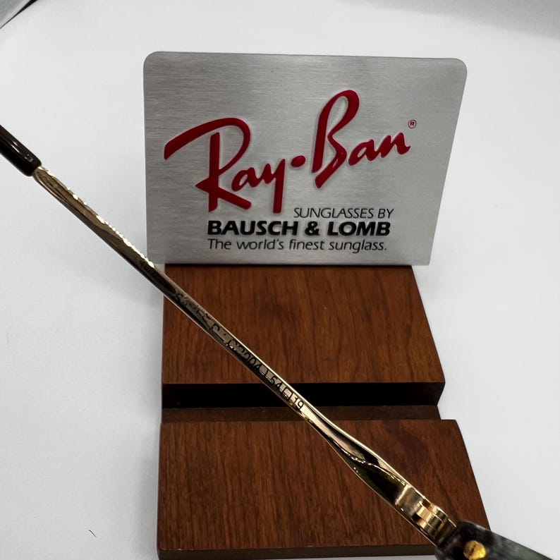 Pu&ograve; includere: Primo piano di occhiali da sole Ray-Ban con montatura argentata e dorata. Il logo Ray-Ban &egrave; rosso su sfondo argentato. Il testo "SUNGLASSES BY BAUSCH & LOMB The world's finest sunglass." &egrave; sotto.