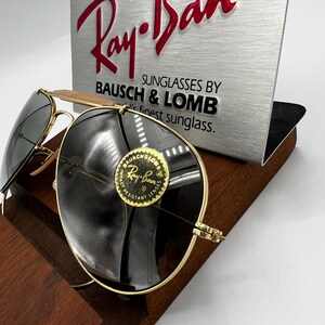 May include: Gold-framed Ray-Ban sunglasses with dark lenses. The sunglasses are displayed on a wooden stand, with a silver sign behind them that reads "Ray-Ban" and "Sunglasses by Bausch & Lomb". A round sticker on the lens says "Ray-Ban".