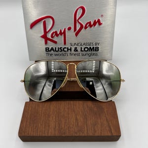 K&ouml;nnte beinhalten: Goldene Aviator-Sonnenbrille mit verspiegelten Gl&auml;sern. Die Sonnenbrille liegt auf einem Holzst&auml;nder. Das Ray-Ban-Logo und der Text "Sunglasses by Bausch & Lomb. The world's finest sunglass." sind auf silbernem Hintergrund sichtbar.