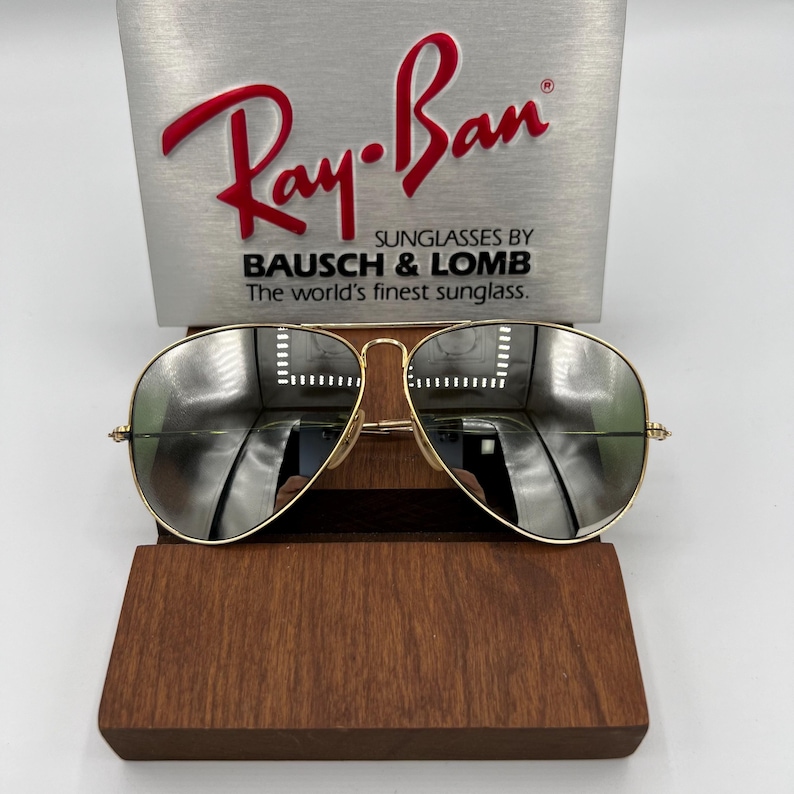 K&ouml;nnte beinhalten: Goldene Aviator-Sonnenbrille mit verspiegelten Gl&auml;sern. Die Sonnenbrille liegt auf einem Holzst&auml;nder. Das Ray-Ban-Logo und der Text "Sunglasses by Bausch & Lomb. The world's finest sunglass." sind auf silbernem Hintergrund sichtbar.