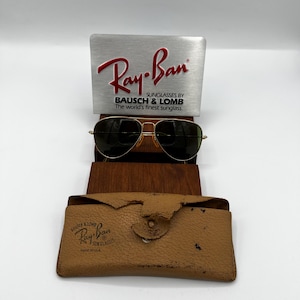 May include: Gold-framed Ray-Ban sunglasses with dark lenses, displayed with a silver sign and a brown leather case. The sign reads "Ray-Ban" and "Sunglasses by Bausch & Lomb". The case is tan and has the Ray-Ban logo.