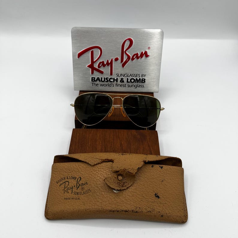 May include: Gold-framed Ray-Ban sunglasses with dark lenses, displayed with a silver sign and a brown leather case. The sign reads "Ray-Ban" and "Sunglasses by Bausch & Lomb". The case is tan and has the Ray-Ban logo.