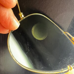 May include: Close-up of gold-rimmed sunglasses with dark lenses. The glasses have a classic aviator shape, with a thin gold frame and a bridge. The lenses are dark, and the glasses are held by a person's fingers.