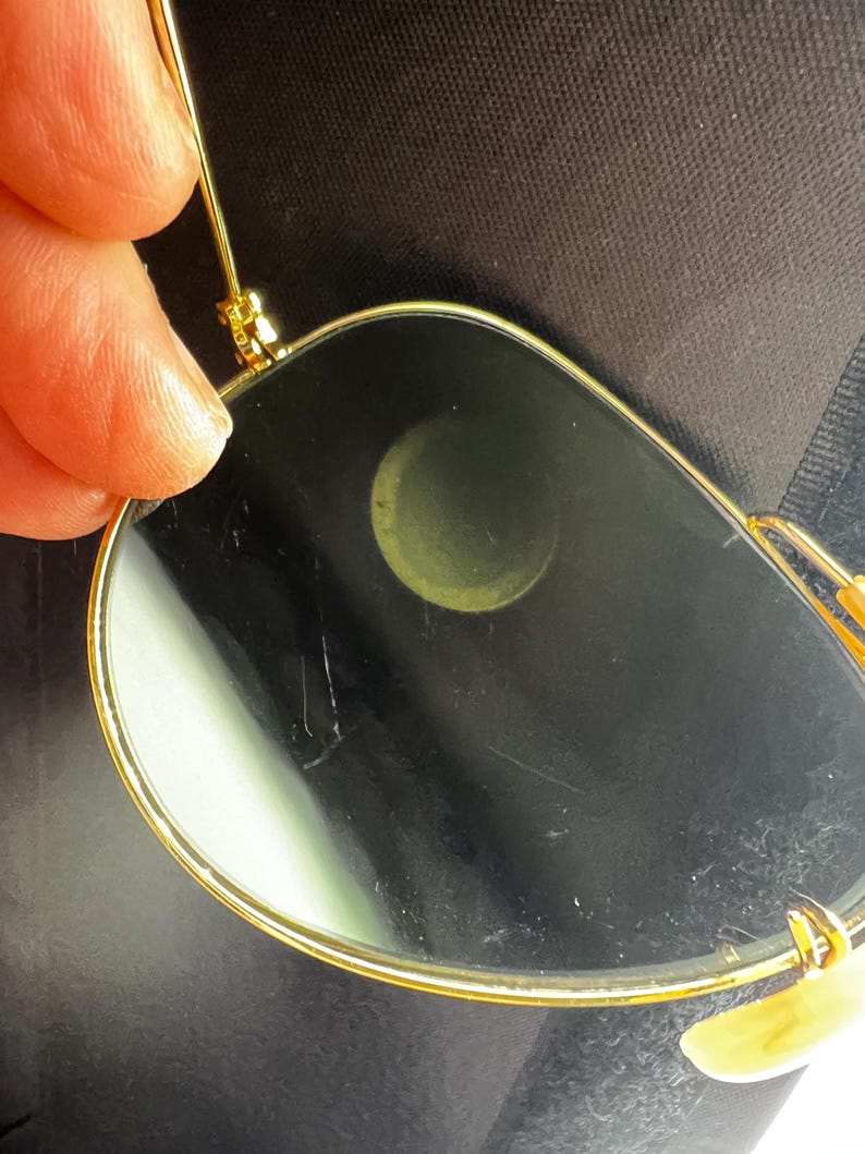 May include: Close-up of gold-rimmed sunglasses with dark lenses. The glasses have a classic aviator shape, with a thin gold frame and a bridge. The lenses are dark, and the glasses are held by a person's fingers.