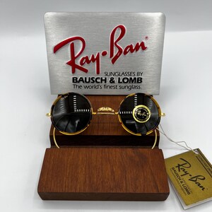 K&ouml;nnte beinhalten: Runde Ray-Ban Sonnenbrille mit goldenem Gestell und dunklen Gl&auml;sern, pr&auml;sentiert auf einem Holzst&auml;nder. Das Ray-Ban-Logo ist auf den Gl&auml;sern und dem St&auml;nder zu sehen. Ein Etikett mit dem Ray-Ban-Logo und Text ist ebenfalls vorhanden.