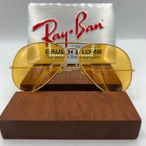 Puede incluir: Gafas de sol tipo aviador con monturas finas de metal dorado y lentes tintadas en amarillo. Las gafas est&aacute;n colocadas frente a un letrero plateado con el logotipo rojo de Ray-Ban y el texto Sunglasses by Bausch & Lomb, The world's finest sunglass.