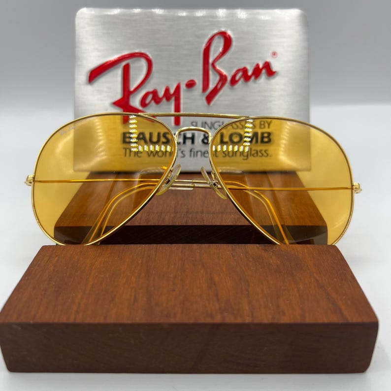 Puede incluir: Gafas de sol tipo aviador con monturas finas de metal dorado y lentes tintadas en amarillo. Las gafas est&aacute;n colocadas frente a un letrero plateado con el logotipo rojo de Ray-Ban y el texto Sunglasses by Bausch & Lomb, The world's finest sunglass.