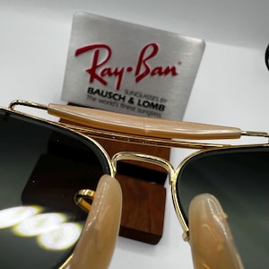 May include: Gold-rimmed sunglasses with dark lenses. The sunglasses have a light brown bridge and are displayed in front of a metal sign with the Ray-Ban logo and text.