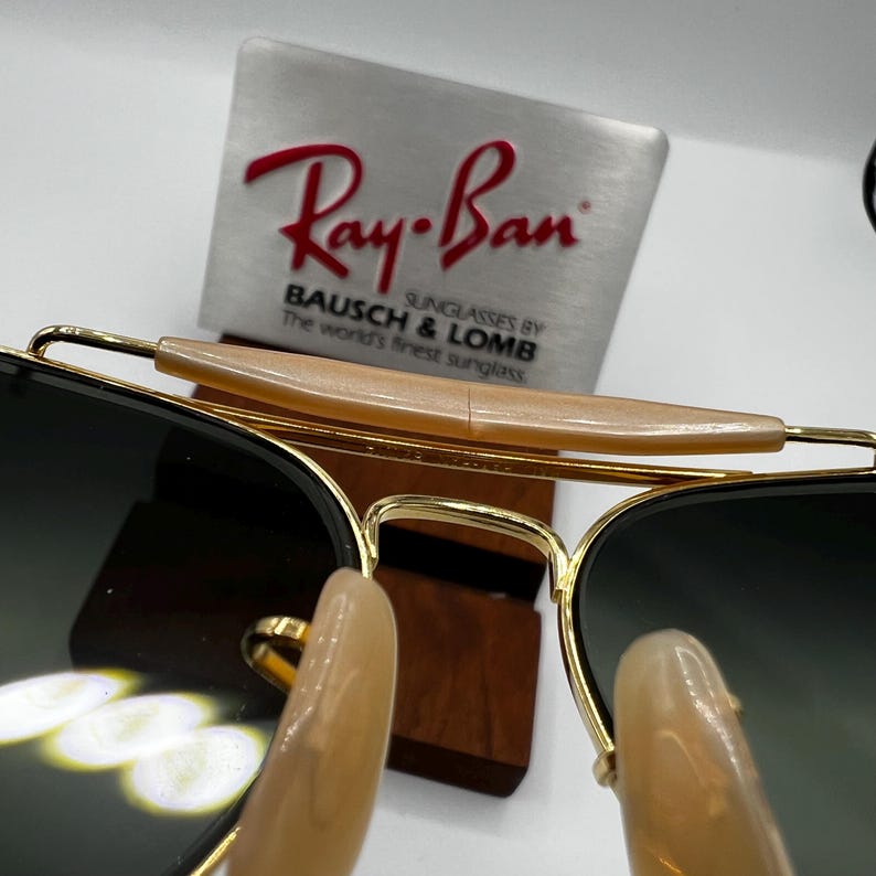 May include: Gold-rimmed sunglasses with dark lenses. The sunglasses have a light brown bridge and are displayed in front of a metal sign with the Ray-Ban logo and text.