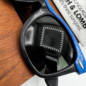Op de afbeelding: Zwarte zonnebril met blauw gemarmerde accenten op de armen. De lenzen reflecteren een vierkant patroon van lichten. De zonnebril ligt op een houten oppervlak. Een wit label met de tekst "SUNGLASSES B & LOMB" is zichtbaar.