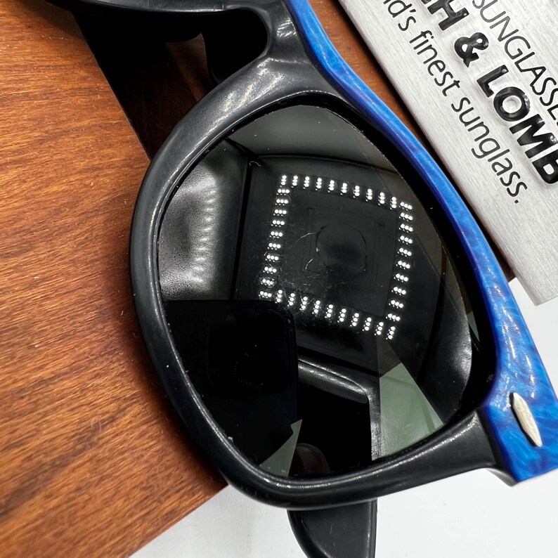 Op de afbeelding: Zwarte zonnebril met blauw gemarmerde accenten op de armen. De lenzen reflecteren een vierkant patroon van lichten. De zonnebril ligt op een houten oppervlak. Een wit label met de tekst "SUNGLASSES B & LOMB" is zichtbaar.