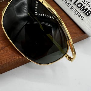 Puede incluir: Gafas de sol con montura dorada y lentes oscuras. Las gafas de sol tienen forma rectangular y una fina montura dorada. Las lentes son de color oscuro s&oacute;lido, y las gafas de sol est&aacute;n sobre una superficie de madera. El texto "finest sunglass" es parcialmente visible.