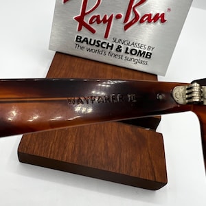 May include: Close-up of Ray-Ban Wayfarer II sunglasses. The frames are a tortoiseshell pattern. The text "WAYFARER II" is visible on the arm. The Ray-Ban logo and "BAUSCH & LOMB" are displayed on a silver sign.