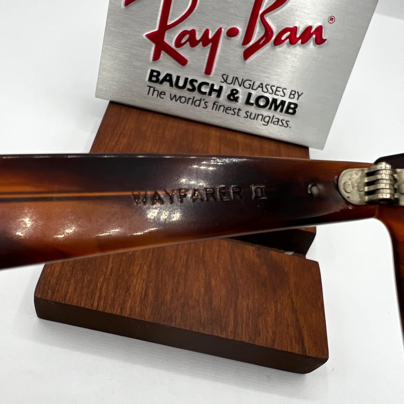May include: Close-up of Ray-Ban Wayfarer II sunglasses. The frames are a tortoiseshell pattern. The text "WAYFARER II" is visible on the arm. The Ray-Ban logo and "BAUSCH & LOMB" are displayed on a silver sign.