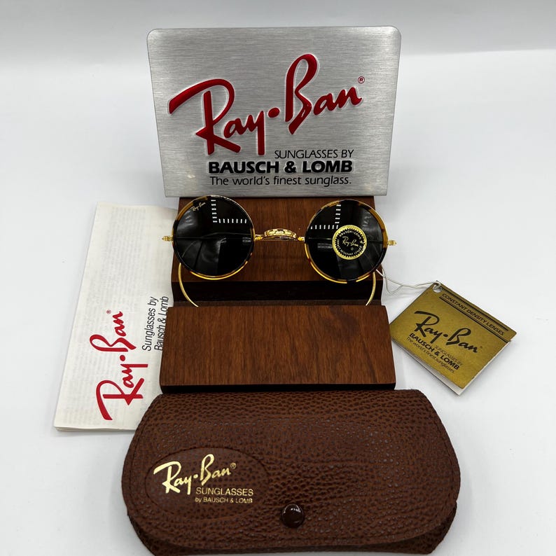 K&ouml;nnte beinhalten: Ray-Ban Sonnenbrille mit schwarzen runden Gl&auml;sern und goldenen Rahmen, pr&auml;sentiert mit einem Holzetui, einem Lederetui und einer Markenverpackung. Das Ray-Ban-Logo ist auf der Verpackung und der Sonnenbrille sichtbar.