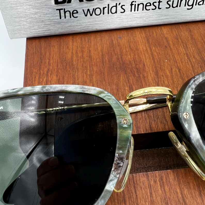 Pu&ograve; includere: Occhiali da sole con montatura marmorizzata verde e grigia e lenti scure. Il ponte e le aste dorate collegano le lenti. Il testo "The world's finest sunglasses" &egrave; visibile sullo sfondo.