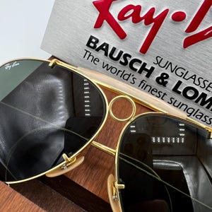 Pode incluir: Close-up de &oacute;culos de sol Ray-Ban com arma&ccedil;&otilde;es douradas e lentes escuras. Os &oacute;culos de sol s&atilde;o exibidos em uma superf&iacute;cie de madeira com um anel dourado e uma placa que diz "Ray-Ban" e "BAUSCH & LOMB SUNGLASSES".