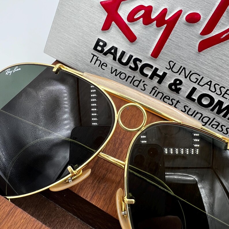 Pode incluir: Close-up de &oacute;culos de sol Ray-Ban com arma&ccedil;&otilde;es douradas e lentes escuras. Os &oacute;culos de sol s&atilde;o exibidos em uma superf&iacute;cie de madeira com um anel dourado e uma placa que diz "Ray-Ban" e "BAUSCH & LOMB SUNGLASSES".