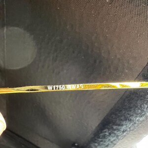 K&ouml;nnte beinhalten: Nahaufnahme eines goldfarbenen Metallrahmens mit dem eingravierten Text "W1750 WWAS". Der Rahmen ist d&uuml;nn und scheint zu einer Brille oder Sonnenbrille zu geh&ouml;ren. Der Hintergrund ist dunkel und unscharf.