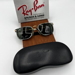 Pu&ograve; includere: Occhiali da sole Ray-Ban con custodia nera. Gli occhiali hanno una montatura verde e nera marmorizzata con dettagli color oro. La custodia &egrave; nera con il logo Ray-Ban in rilievo. Un cartello argentato con il logo Ray-Ban &egrave; sullo sfondo.