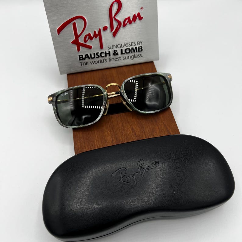 Pu&ograve; includere: Occhiali da sole Ray-Ban con custodia nera. Gli occhiali hanno una montatura verde e nera marmorizzata con dettagli color oro. La custodia &egrave; nera con il logo Ray-Ban in rilievo. Un cartello argentato con il logo Ray-Ban &egrave; sullo sfondo.