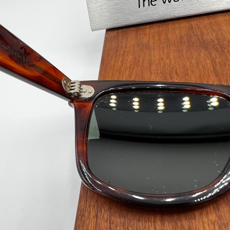 Puede incluir: Primer plano de gafas de sol carey con lentes oscuras. La montura es de color marr&oacute;n c&aacute;lido con un patr&oacute;n moteado. La bisagra es plateada. Las gafas de sol est&aacute;n sobre una superficie de madera.