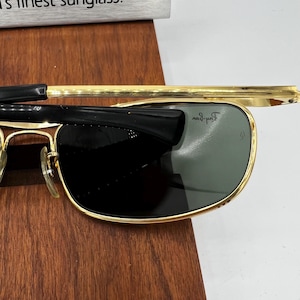 Puede incluir: Gafas de sol con montura dorada, patillas negras y lentes verde oscuro. Las gafas de sol est&aacute;n sobre una superficie de madera. Las lentes tienen una ligera curvatura y la montura es rectangular.